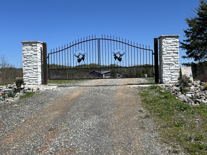 Black iron driveway gate between stone columns at the end of a gravel drive